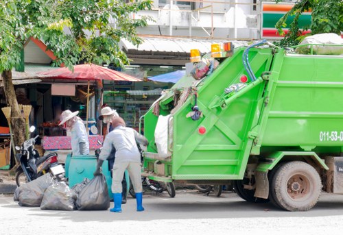 A bright green waste collection truck parked on a street in Greenwich, identifiable by its colour and design, with its rear hatch open, revealing compartments filled with waste materials. In the foreground, two workers wearing light grey uniforms, protective gloves, and caps are engaged in rubbish collection; one is lifting a large black trash bag from the ground, while the other stands nearby. Several black garbage bags are placed on the pavement in front of them, suggesting they are in the process of loading waste into the truck. The background shows a busy urban scene with small shops, awnings, and trees lining the street, typical of a Greenwich locality. The environment appears well-lit, indicative of daytime, with surrounding buildings and shopfronts creating a typical setting for rubbish removal services in the area. Business Waste Removal Greenwich provides professional waste collection within the local South East London postcode area, including Greenwich and nearby districts.