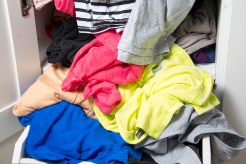 A close-up view of an open white metal drawer filled with a disorganized pile of mixed laundry, including various coloured clothing items such as a bright pink garment, a yellow piece, grey fabric, and blue fabric, with some clothes visibly wrinkled and overlapping each other, set against a background that appears to be part of a domestic storage or laundry area, emphasizing clutter that could be targeted for rubbish removal or clearance services in Greenwich or nearby areas, as offered by Business Waste Removal Greenwich.