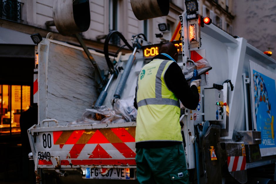 A street cleaner dressed in high-visibility orange uniform with reflective silver stripes, wearing a matching orange cap, is standing on a paved sidewalk next to a concrete wall. The individual is hol