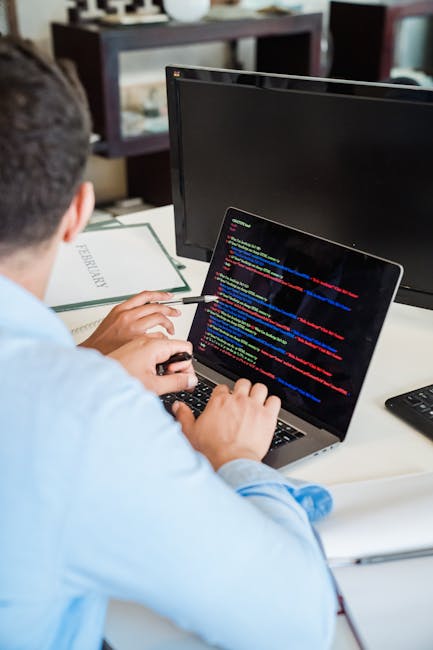 A person with light skin and short fingernails is using a silver MacBook laptop with a black keyboard, placed on a dark, reflective surface. The person's hands are visible, with one hand resting on th