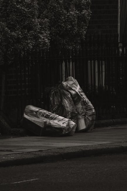 Two large black plastic rubbish bags, slightly crumpled and tied at the top, are positioned on the pavement beside a dark wooden fence. The bags contain mixed waste materials, with some visible white 